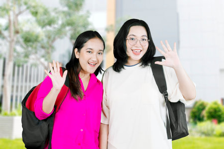 Cheerful teenage girls wearing eyeglasses and school uniform smiling while waving hand at cameraの写真素材