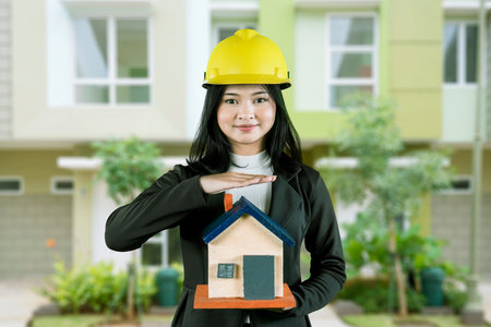 Female Architect Holding a House Model with a Yellow Hard Hat in Front of Residential Buildingsの写真素材