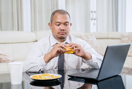 Businessman Eating a Cheeseburger While Working on Laptop at Home â Focused Expressionの写真素材