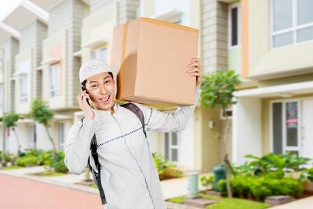 Happy Muslim Delivery Man Holding a Box and Talking on the Phone in a Residential Neighborhoodの写真素材
