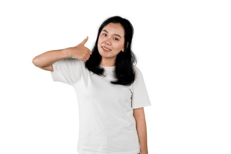Smiling Asian woman giving thumbs up gesture while standing against white backgroundの写真素材
