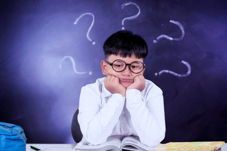 Confused Schoolboy Sitting at Desk with Books and Question Marks on Chalkboard Backgroundの写真素材