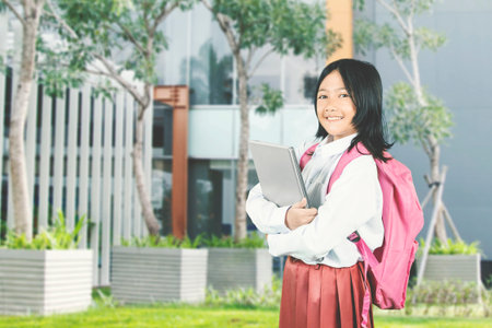 Happy Asian Schoolgirl with Backpack Holding Laptop Outdoorsの写真素材
