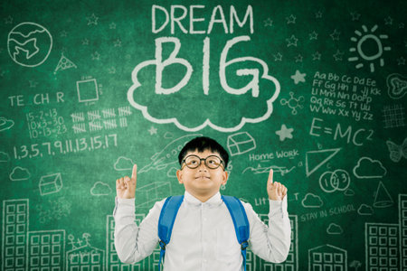 Cheerful Schoolboy with Backpack and Glasses Pointing Up in Front of Inspirational Dream Big...