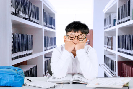 Bored Schoolboy Sitting at Desk in Library with Open Books and Backpack