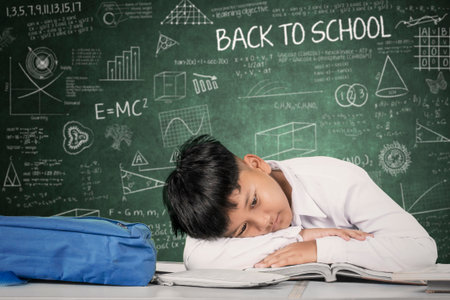 Tired Student Lying on Desk with Head on Book in Front of Chalkboardの写真素材