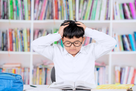 Stressed Schoolboy Studying in Library with Hands on Head and Open Books on Deskの写真素材