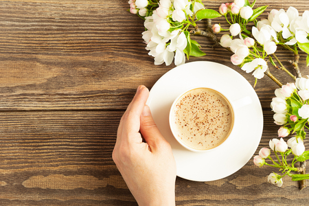 A cup of hot aromatic coffee in female hands and a blooming apple tree branch on a wooden background. Flat layout.の写真素材