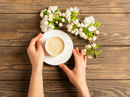 A cup of hot aromatic coffee in female hands and a blooming apple tree branch on a wooden background. Flat layout.の写真素材