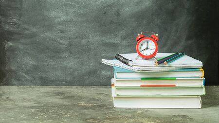 A stack of educational notebooks of books and a red alarm clock on the background of a chalk board....
