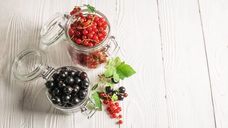 Ripe fresh black and red currant berries in glass jars on white wooden background. Horizontal frame. Selective focus.の写真素材