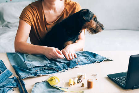 A woman with pet in her arms sews clothes for dog and watches lesson online laptop. DIY Reuse Recycling Zero Wasteの写真素材
