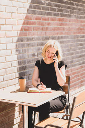 A young woman is talking on cell phone and making notes in a notebook at a table in cafe on city...
