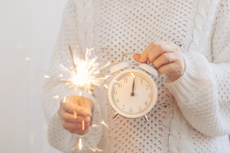 A girl in white sweater holds sparklers, clock is an alarm clock symbol of the new year, arrows at midnight 12 oclock.の写真素材