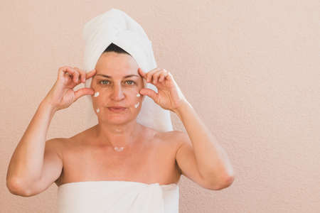 Middle-aged woman with a towel on her head, no makeup, applies cream to her face and makes massage. Facial skin careの写真素材