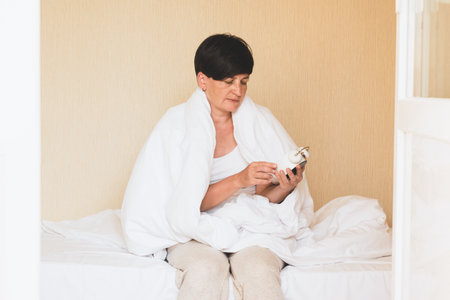 A middle-aged woman is sitting in bed with an alarm clock in her hands. Morning awakening, sleep problems insomniaの写真素材