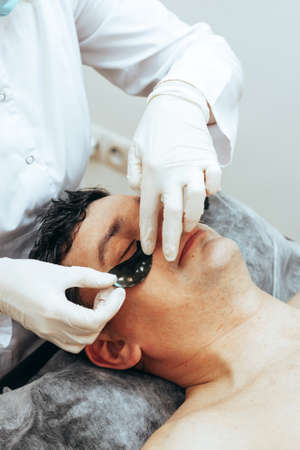 Doctor Cosmetologist applies black patches on eyes of man spa beauty salon. The concept face care for menの写真素材