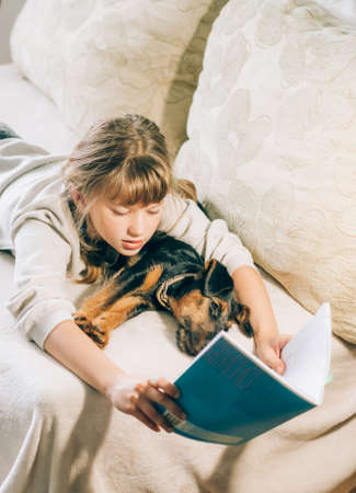A girl and small dog puppy lie on the couch engaged in reading training. The concept of friendship...