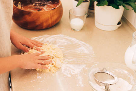 The hands of a young girl are kneading dough for baking on kitchen table. Daily household chores in...