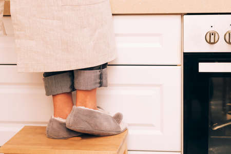 Lovely girls feet in soft slippers stand on wooden stool in a cozy kitchen. The present moment of lifeの写真素材