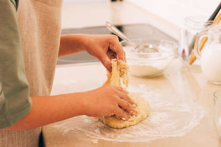 The hands of a young girl are kneading dough for baking on kitchen table. Daily household chores in...