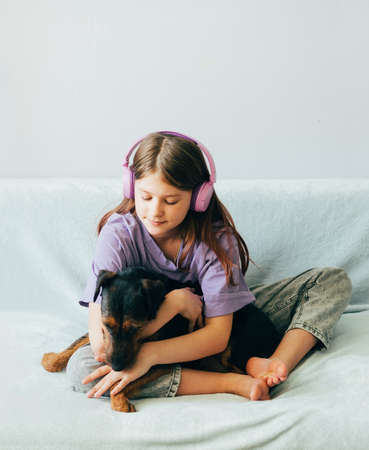 Happy teenage girl in lilac t-shirt in pink headphones listens to music and has fun playing with dog