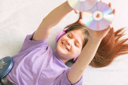 A happy teenage girl in a lilac T-shirt lies on the bed headphones and listens to music on CD playerの写真素材