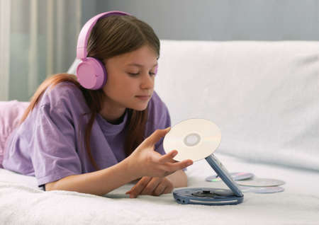 A teenage girl in a lilac T-shirt lies on a bed in pink headphones and listens to music on CD diskの写真素材
