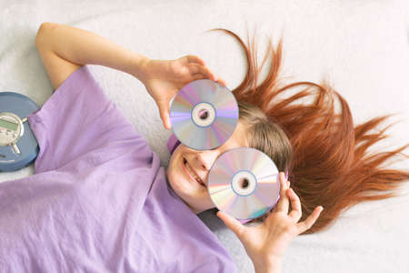 Happy teenage girl in lilac t-shirt lies on bed in pink headphones holds cd disks in her hands