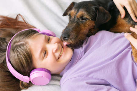 Happy teenage girl in lilac t-shirt in pink headphones listens to music and has fun playing with dogの写真素材