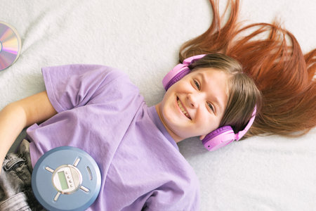 Happy teenage girl in lilac t-shirt lies on bed in pink headphones listens to music on CD playerの写真素材
