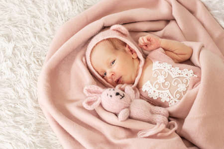 A cute newborn baby girl in a beautiful knitted hat lies under soft blanket with favorite toy hare