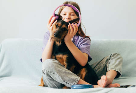 Happy teenage girl in lilac t-shirt in pink headphones listens to music and has fun playing with dog