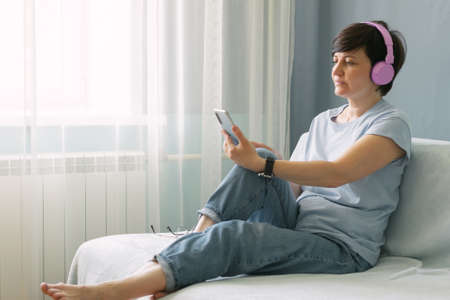 An adult woman in pink headphones sits on the sofa at home and listens to musicの写真素材