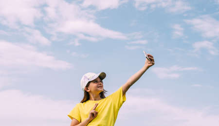 A preteen girl takes a selfie on a mobile phone against a blue sky. Horizontal frame, copy spaceの写真素材