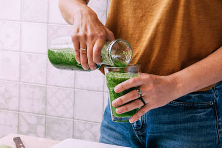A woman pours a freshly prepared smoothie from green vegetables and fruits into glass. Vegetarianism