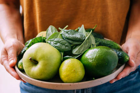 A woman holds in her hands fresh green vegetables and fruits in a large plate, close-up soft focusの写真素材