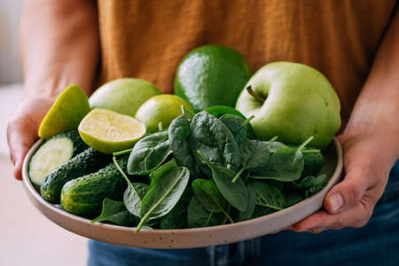A woman holds in her hands fresh green vegetables and fruits in a large plate, close-up soft focusの写真素材