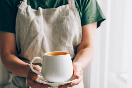 A Barista man or waiter in a linen apron serves white cup of coffee on a saucer. Hot drink in handsの写真素材