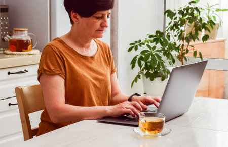 Mature woman in casual clothes works on laptop at a wooden table with a cup of tea, surrounded by indoor plants in a cozy kitchen settingの写真素材