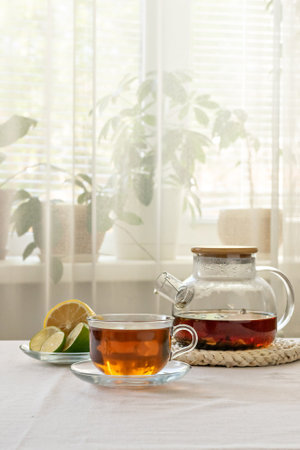 Glass teapot with brewed tea, cup of tea, slices of lemon and lime and sugar cubes on the table against the background of a light window with light curtains, vertical frameの写真素材