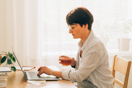 Mature woman holding a cup of tea and working on a laptop at a wooden table, surrounded by indoor plants in a bright roomの写真素材