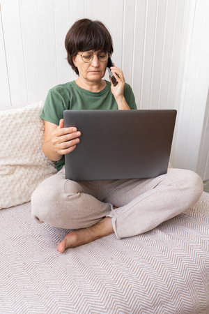 Mature woman using laptop and phone on sofa in relaxed home settingの写真素材