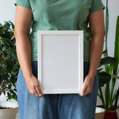 Woman in green t-shirt and blue jeans holding empty frame in space filled with plants against white wall. Modern minimalist designの写真素材