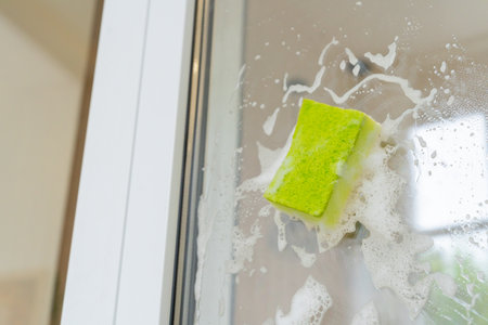 Green sponge cleaning glass shower door with soap suds in bathroom
