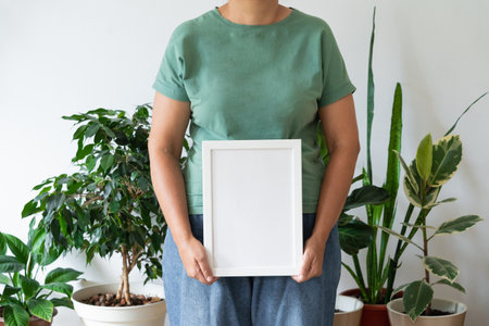 Woman in green t-shirt and blue jeans holding empty frame in space filled with plants against white...