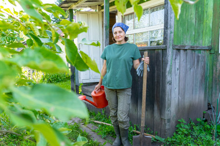 Caucasian mature female gardener with watering can and shovel in rural garden setting. Eco-friendly lifestyle, natural farmingの写真素材