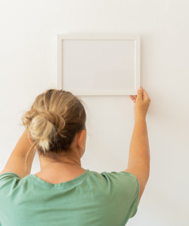 Blonde woman in green t-shirt hanging empty picture frame on white wall at home. Modern minimalist...