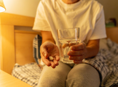 A mature woman sits on her bed at night holding pills and a glass of water. Sleep problems, depression, psychological disorders, addictionの写真素材