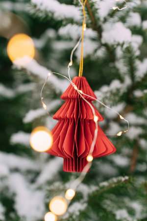 Red paper Christmas tree ornament hanging on snowy pine with lights. Festive composition, mockup for a New Years cardの写真素材